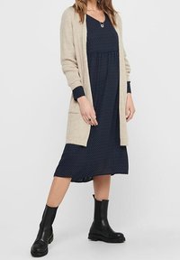 Beige pletený cardigan přes tmavě modré šaty s puntíky, s volným střihem a dlouhými rukávy. Černé kotníkové boty dotvářejí celý outfit.