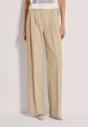 Beige broek met wijde pijpen en een gestructureerde afwerking, voorzien van plooien aan de voorkant en een tailleband op halverwege de heup. Gecombineerd met lichtgekleurde puntige schoenen.