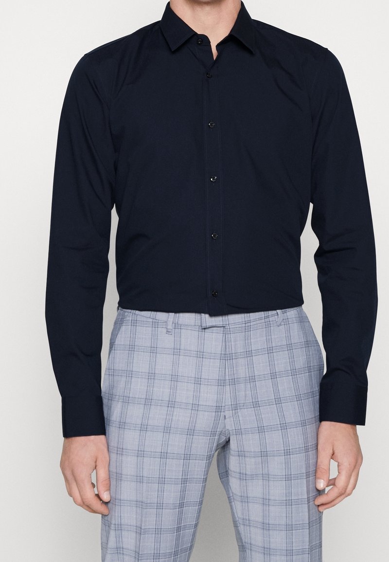 Chemise bleu marine à boutons, avec un col structuré et des manches longues, fabriquée en tissu lisse, associée à un pantalon à carreaux gris clair.