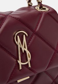 Bolso de mano de imitación piel burdeos con diseño acolchado, que presenta herrajes en tono dorado y detalles cosidos. Logotipo exhibido de forma destacada en la parte frontal.