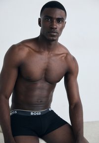 Schwarze Boxershorts mit einem grauen Bund, auf dem das "BOSS"-Logo zu sehen ist. Das Modell hat einen muskulösen Körperbau und kurzes, strukturiertes Haar.
