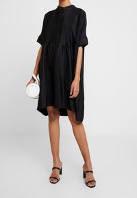 Abito nero con maniche corte, silhouette rilassata e chiusura frontale con bottoni. Abbinato a una borsa bianca circolare e sandali neri con tacco a blocco.
