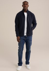 Tumed sinine fliistek jope kõrge kaelusega, ees lukuga tasku, kantud valge T-särgi peal, kombineeritud siniste teksade ja valgete tossudega.