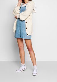 Kardigan yang terbuat dari rajutan krim dengan kancing besar dilapisi di atas gaun overall denim. Sneakers putih dengan model tinggi melengkapi penampilan.