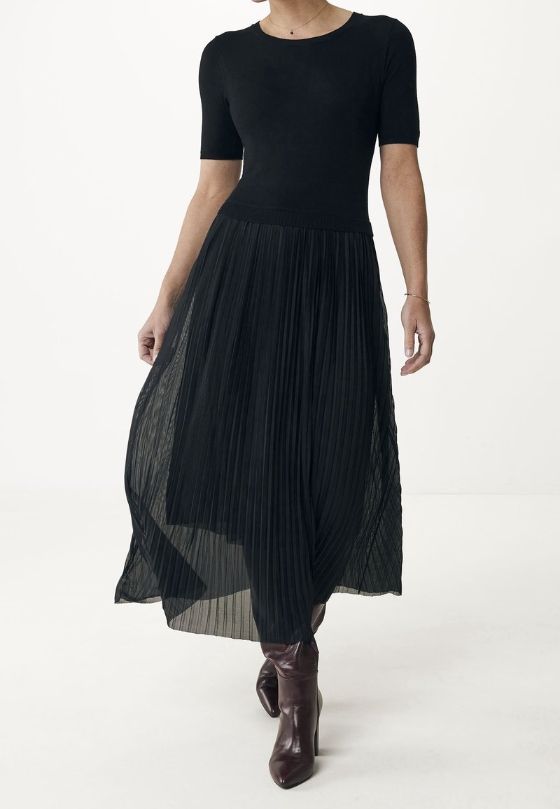 Černé šaty s těsným korzetem s krátkým rukávem a skládacím, průhledným sukní, doplněné lesklými vínovými kozačkami po kolena.