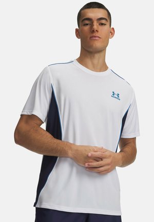 Mann trägt ein weißes Sport-T-Shirt mit dunkelblauen Seitenpaneelen und einem blauen Under Armour-Logo auf der Brust, steht mit verschränkten Händen.
