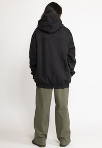 Zwarte hoodie met capuchon, voorzien van een textuurontwerp van bergen in zwarte draad op de achterkant, gecombineerd met losse olijfgroene broek.
