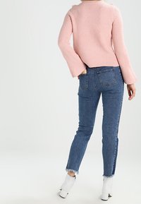 Personne portant un pull côtelé rose, un jean taille haute à ourlet effiloché bleu, et des bottines blanches, debout avec une main dans la poche.