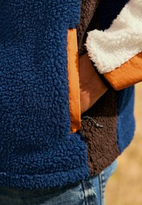 Fleecejas met blauwe, bruine en witte delen, voorzien van een oranje voering en een elastische zoom. Inclusief koordverstelling in de taille.