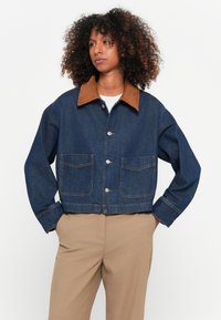 Afgekorte spijkerjack in donkerblauw met een bruine suede kraag. Voorzien van twee voorzakken en zilveren knoopsluitingen. Zachte textuur.