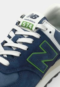 Blue suede sneakers featuring white laces and a green accent on the signature logo. Model number "574" displayed on the tongue.