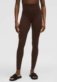 Leggings marron taille haute en tissu extensible et lisse ; conception ajustée avec une taille sans couture. Associés à des sandales noires à enfiler.