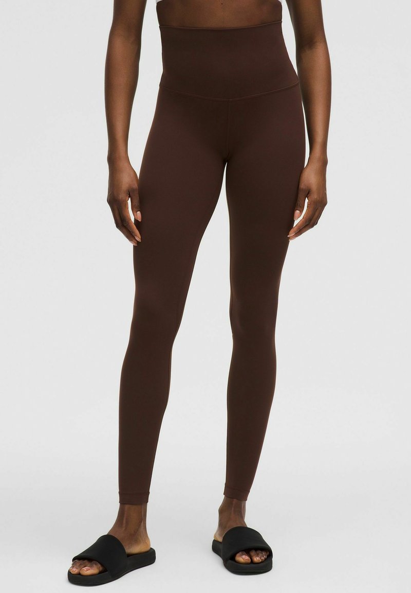 Leggings marron taille haute en tissu extensible et lisse ; conception ajustée avec une taille sans couture. Associés à des sandales noires à enfiler.