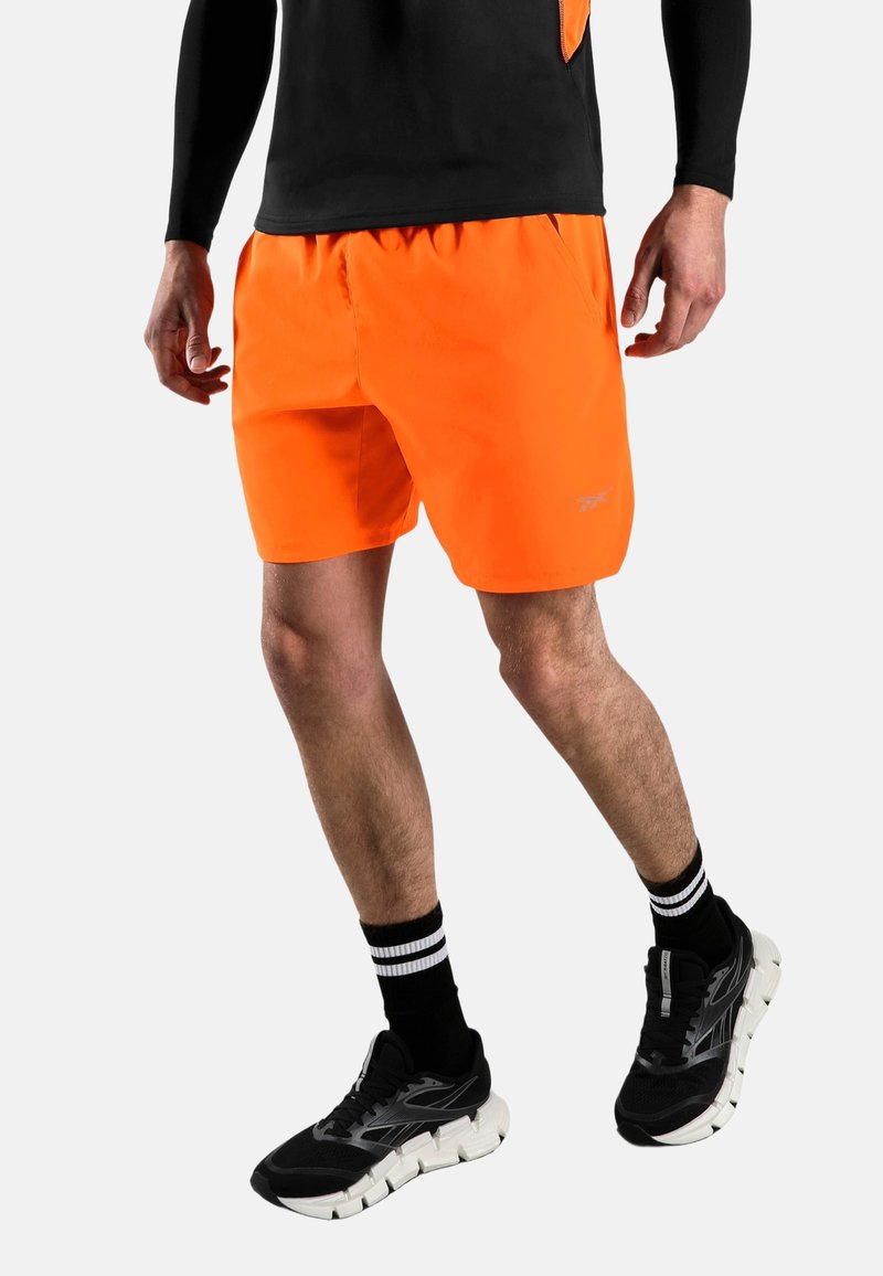 Homme portant un short de sport orange vif, un haut à manches longues noir, des chaussettes rayées noires et blanches, et des chaussures de course noires sur fond blanc.