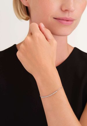 Zilveren armband met heldere edelstenen, gezet in een delicate ketting, die een gebogen ontwerp toont tegen een donkere achtergrond.