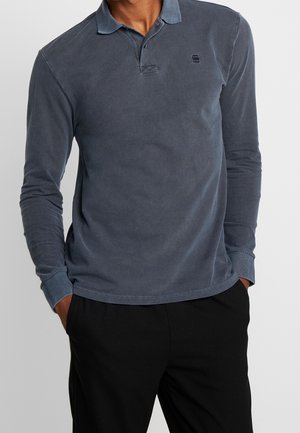 Mężczyzna w długim rękawie, wyblakłej niebieskiej koszulce polo z kołnierzykiem i czarnych spodniach, z rękami w kieszeniach, na jednolitym tle.