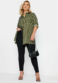 Camicia a righe verde e bianca con bottoni, colletto e taschino, abbinata a pantaloni neri slim-fit e una piccola borsa nera.