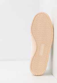 Brązowa gumowa podeszwa z teksturowanymi wzorami; minimalistyczna kremowa cholewka z widocznymi szwami; widok pod kątem na jednolitym tle.