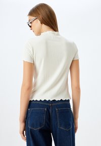 Kortærmet ribbet top i off-white med scalloped kant og en subtil tekstur. Bæres med mørkeblå højtaljede jeans.