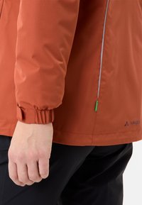 Giacca arancione leggera con tessuto strutturato, polsini elasticizzati e dettagli riflettenti. Presenta un'etichetta logo verde e dettagli nelle cuciture laterali.