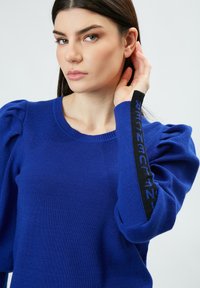 Königsblauer Strickpullover mit Puffärmeln und gerippter Textur; verfügt über einen schwarzen Akzent am Ärmel mit Textdetail.