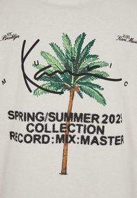 Hvit bomull T-skjorte med grafikk av palmetre, svart tekst med "Karl Kani Music" og "Vår/Sommer 2023-kolleksjon," i varierende skrifttyper.