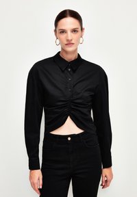 Sort langærmet skjorte med knaplukning foran og en rynket design ved taljen. Lavet af glat stof, parret med tætsiddende sorte jeans.