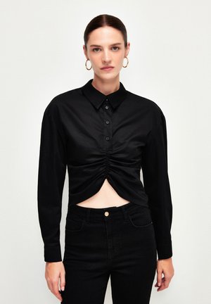 Schwarzes Langarmhemd mit knöpfiger Front und gerafftem Design an der Taille. Aus glattem Stoff gefertigt, kombiniert mit eng anliegenden schwarzen Jeans.