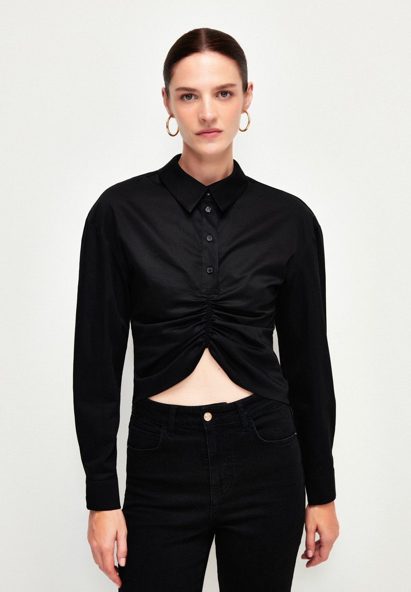 Sort langærmet skjorte med knaplukning foran og en rynket design ved taljen. Lavet af glat stof, parret med tætsiddende sorte jeans.