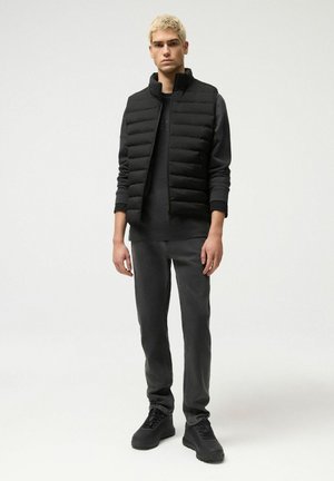 Młody mężczyzna o krótkich blond włosach, ubrany w czarną kamizelkę puchową, długi rękaw koszuli, ciemne jeansy i czarne trampki, stoi na tle jednolitego tła.