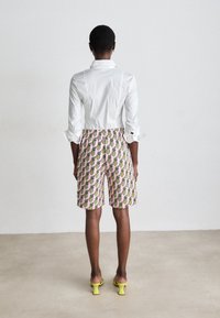 Nainen seisoo selkä kameraan päin valkoisessa paidassa, vaaleanpunaisissa shortseissa, joissa on keltaista ananaskuviota, ja keltaisissa korkokengissä yksinkertaisella lattialla ja seinällä.