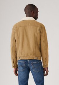 Chaqueta de denim color beige con un cuello de borrego blanco, que presenta un diseño clásico de camionero, botones de metal y detalles en la costura trasera.