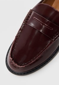 Chaussure penny loafer en cuir brun foncé poli avec surpiqûres visibles et bord de semelle noir, photographiée vue de dessus.