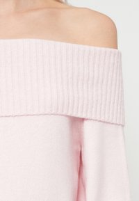 Rozā džemperis ar atklātu plecu, ar ribotu apkakli un mīkstu, teksturētu materiālu. Dizains ir brīvas formas ar gludu apdari.