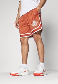 Pantalones cortos de baloncesto de tela de malla naranja, con ribete blanco y un logotipo gráfico de "World Series" en la pierna izquierda. Llevados con zapatillas deportivas blancas.