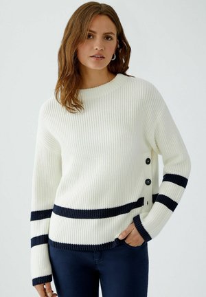 Cremepullover aus Rippstrickstoff mit navyblauen horizontalen Streifen und Knopfdetails an der Seite.