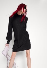 Robe noire pailletée avec un col montant, des manches ballon et une coupe décontractée. Accompagnée d'une pochette argentée ornée de franges.