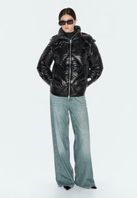 Giacca puffa nera con finitura lucida, dotata di cappuccio, design trapuntato diagonale e zip frontale. Abbinata a jeans a gamba larga blu chiaro.