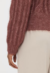 Πλεκτό πουλόβερ σε βαθύ μπορντό, με ανάγλυφη υφή και ελαφρώς φαρδιά μανίκια. Συνδυασμένο με �ανοιχτό μπεζ παντελόνι.