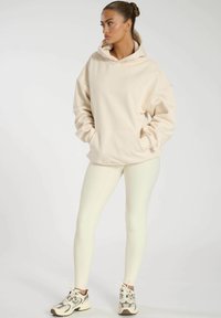 Sweatshirt oversize crème avec poche avant, associé à des leggings blancs à côtes et à des sneakers neutres avec des accents et des lacets.