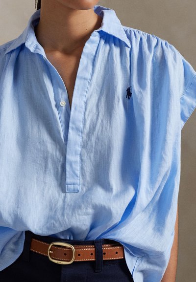 Sieviete, kas valkā gaiši zilu krekla kreklu ar melnu izšūtu logo, ieliektu tumši zilās biksēs ar brūnu ādas jostu ar zelta sprādzi.