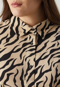 Smėlinis marškinėlis su juodu tigro juostelių raštu, tekstūruota medžiaga ir klasikinis apykaklė su juodu mygtukų uždarymu.