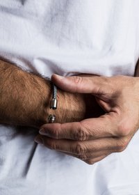 Braccialetto in tessuto grigio intrecciato con accenti metallici, dotato di una chiusura cilindrica. Mano che tiene il braccialetto su un avambraccio.
