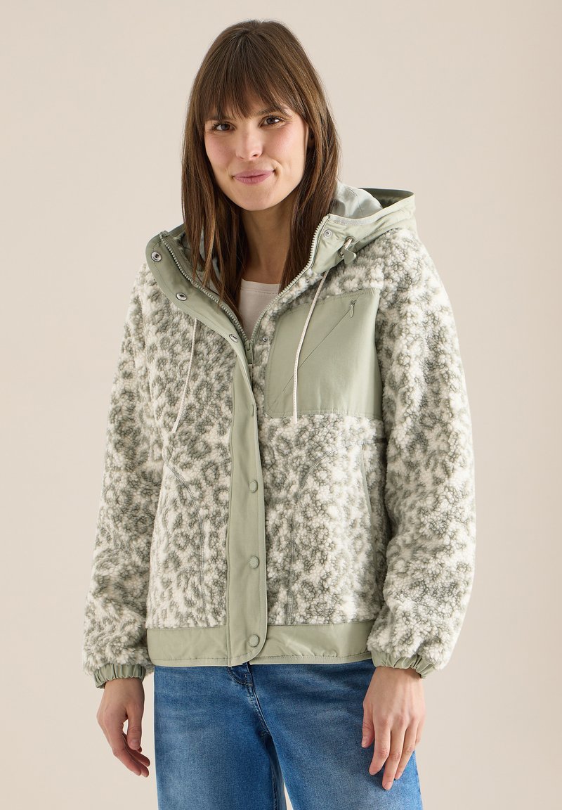 Femme portant une veste polaire à motif vert clair et blanc avec capuche et fermeture éclair à l'avant, associée à un jean bleu, sur un fond uni.