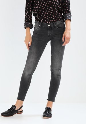 Vrouw die een zwart patroon langharig shirt draagt, vervaagde zwarte skinny jeans met onafgewerkte zoom en zwarte veter-slingbackschoenen.