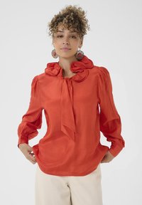 Blouse rouge avec un grand col froncé et un détail à nouer, fabriquée à partir d'un tissu lisse. Présente des manches bouffantes et des fronces aux épaules.
