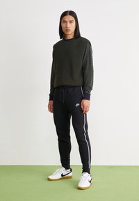 Zeleni rebrasti pleteni bezrukavnik preko tamnoplavog majice dugih rukava sa prugama, u kombinaciji s crnim sportskim hlačama i bijelim tenisicama s naglascima.