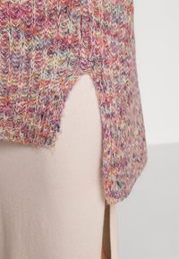 Détail latéral d'un pull tricoté multicolore avec une fente courbée dévoilant une jupe beige en dessous.