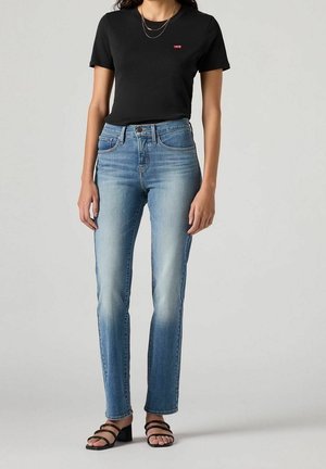 Vrouw met een zwart Levi's T-shirt, lichtblauwe high-waisted jeans, zwarte sandalen met blokhak en open tenen, en gelaagde gouden kettingen.