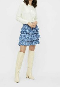 Chemise blanche à col boutonné, jupe à volants bleue à motifs, et bottes en cuir beige montantes jusqu'au genou avec talons carrés. Détails en tissu texturé.